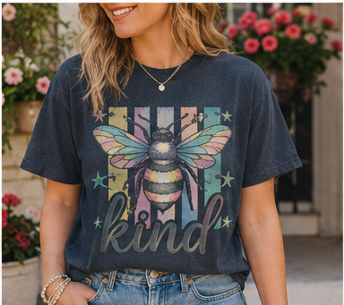 A woman wearing a blue t-shirt with a colorful graphic of a bee and the word "kind" printed on it, standing in front of a window with pink flowers.