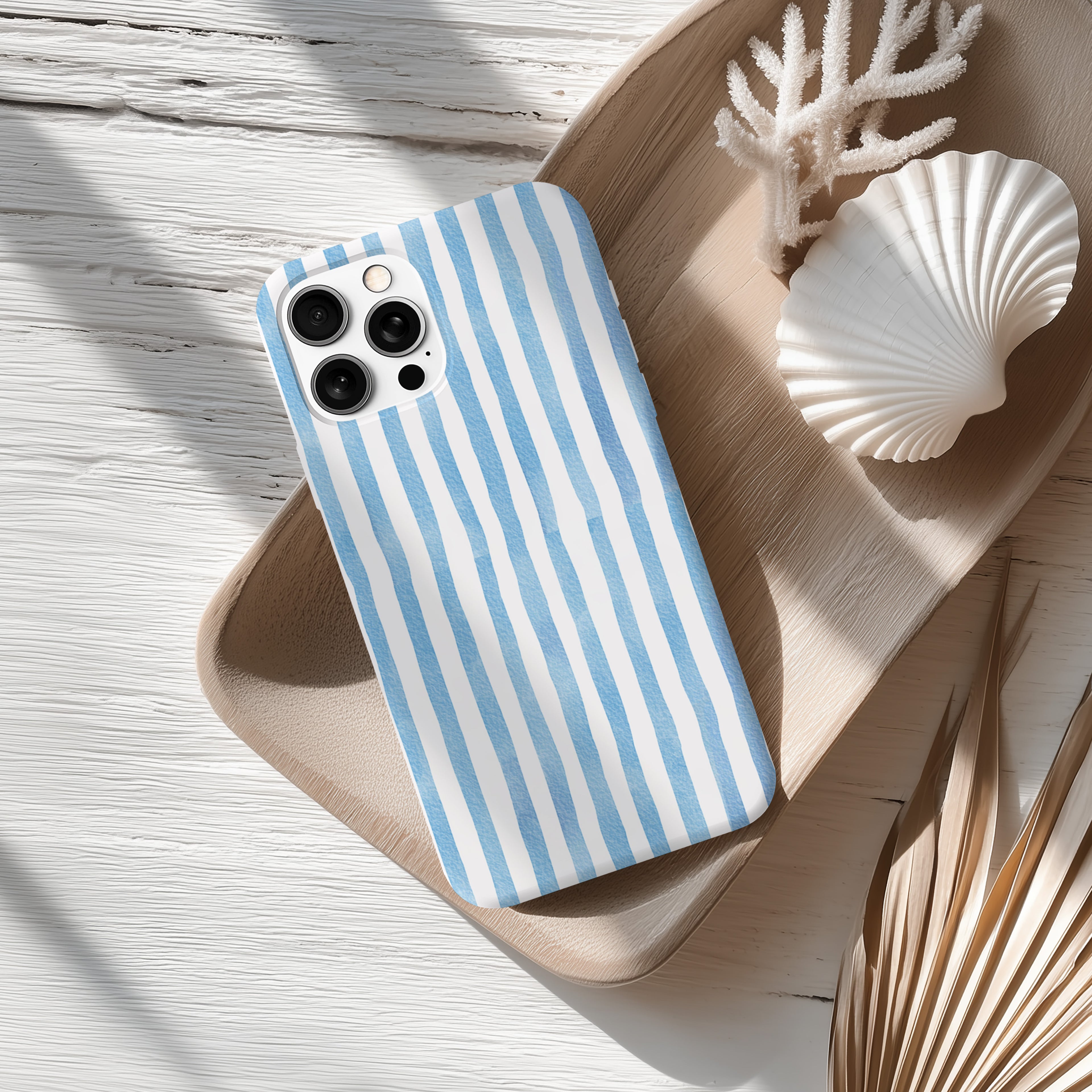 A blue and white striped phone case rests on a wooden surface, accompanied by a white seashell and some dried leaves.