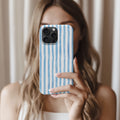 A woman with long, wavy blonde hair is holding a smartphone with a blue and white striped patterned case, taking a selfie.
