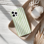 A green and white striped phone case is placed on a wooden surface, accompanied by a seashell and some dried leaves.