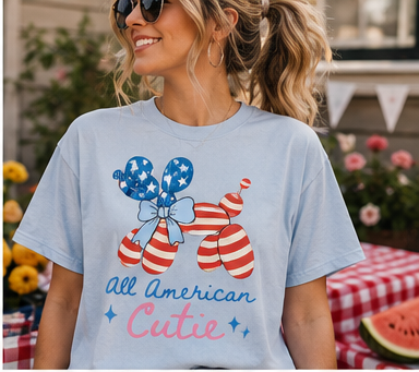 A woman wearing a t-shirt with an American flag design and the text "All American Cutie" is smiling and posing in front of a table with a red and white checkered tablecloth.