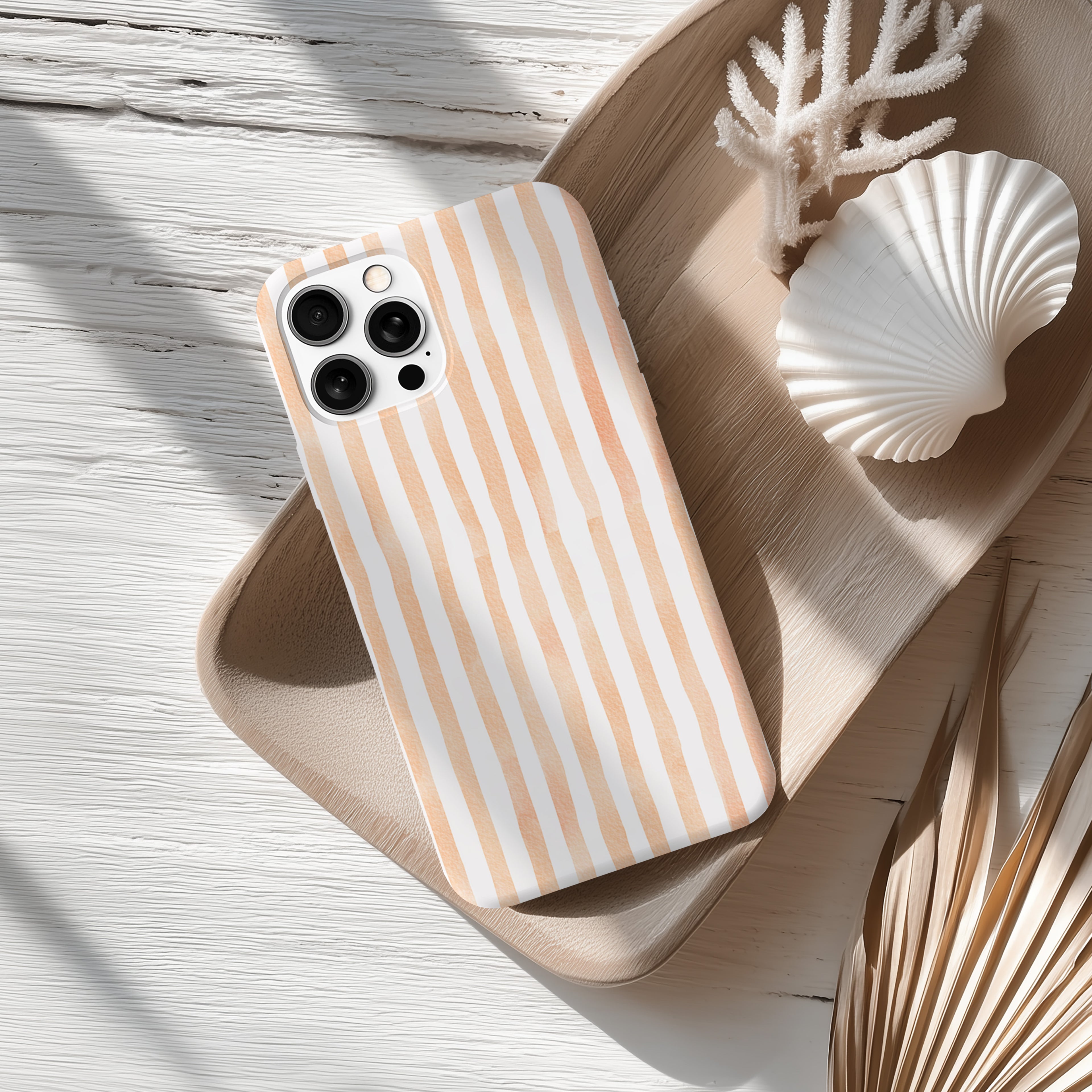 A striped phone case with a light orange and white pattern rests on a wooden surface, accompanied by a seashell and some dried leaves.