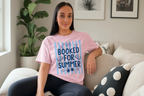 A young woman wearing a pink t-shirt with the text "BOOKED FOR SUMMER" is sitting on a couch in a living room.