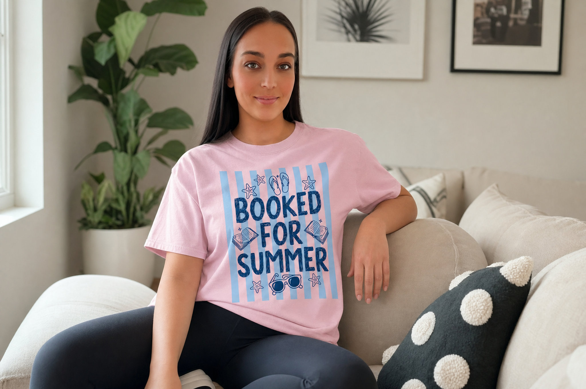 A young woman wearing a pink t-shirt with the text "BOOKED FOR SUMMER" is sitting on a couch in a living room.