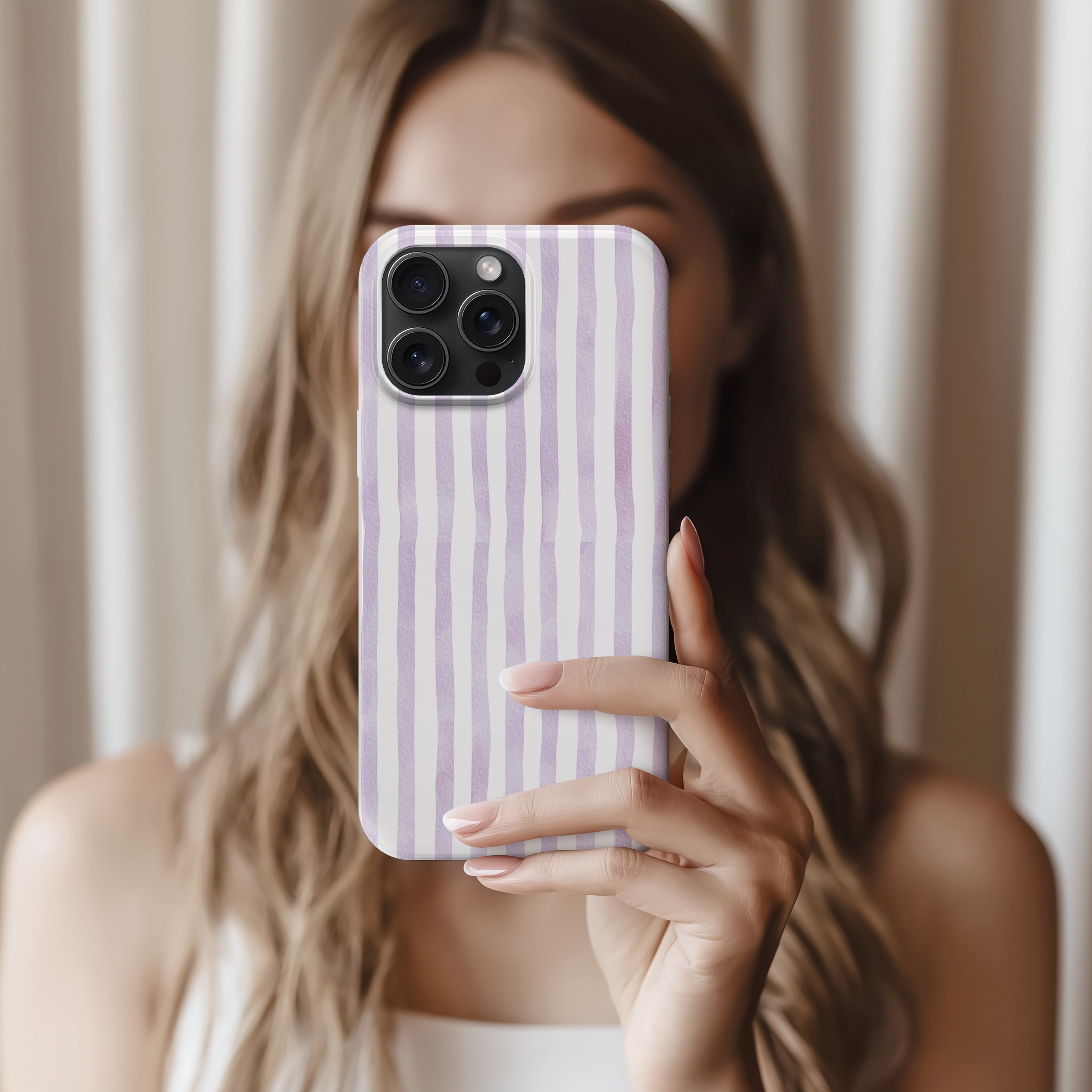 A woman with long, wavy blonde hair is holding a smartphone with a purple and white striped patterned case, taking a selfie.