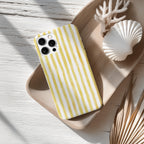 A yellow and white striped phone case is placed on a wooden surface, accompanied by a white seashell and some dried leaves.