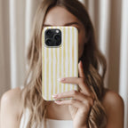 A woman with long, wavy blonde hair is holding a phone with a yellow and white striped pattern, taking a selfie in front of a white curtain.