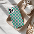 a checkered phone case with a pink and green pattern, placed on a wooden surface alongside a white seashell and some dried leaves.