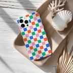 A colorful checkered patterned phone case is placed on a wooden surface, accompanied by a white seashell and some dried leaves.