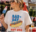 A woman wearing sunglasses and a red bandana is holding a popsicle and smiling, with a white t-shirt featuring a graphic of a hot dog and the text "Bad day to be a hot dog".