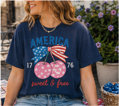 A woman wearing a blue t-shirt with a graphic design of cherries and an American flag, sitting in front of a potted plant with purple flowers.