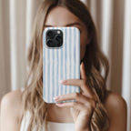 A woman with long, wavy blonde hair is holding a smartphone with a blue and white striped patterned case, taking a selfie in front of a white curtain.