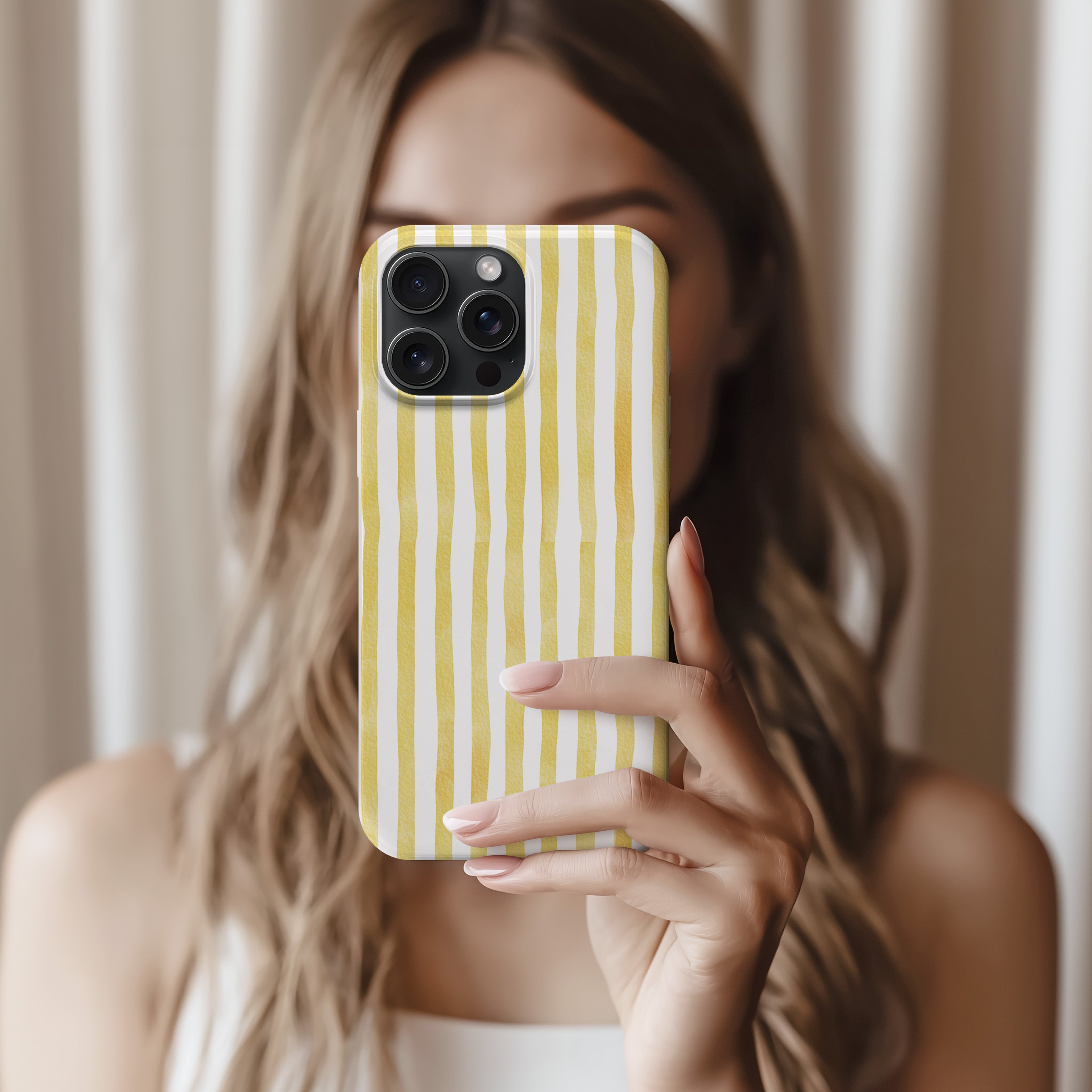 A woman with long, wavy blonde hair is holding a phone with a yellow and white striped pattern, taking a selfie in front of a white curtain.