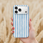 A hand holding a smartphone with a blue and white striped patterned case, against a blurred background of a field.