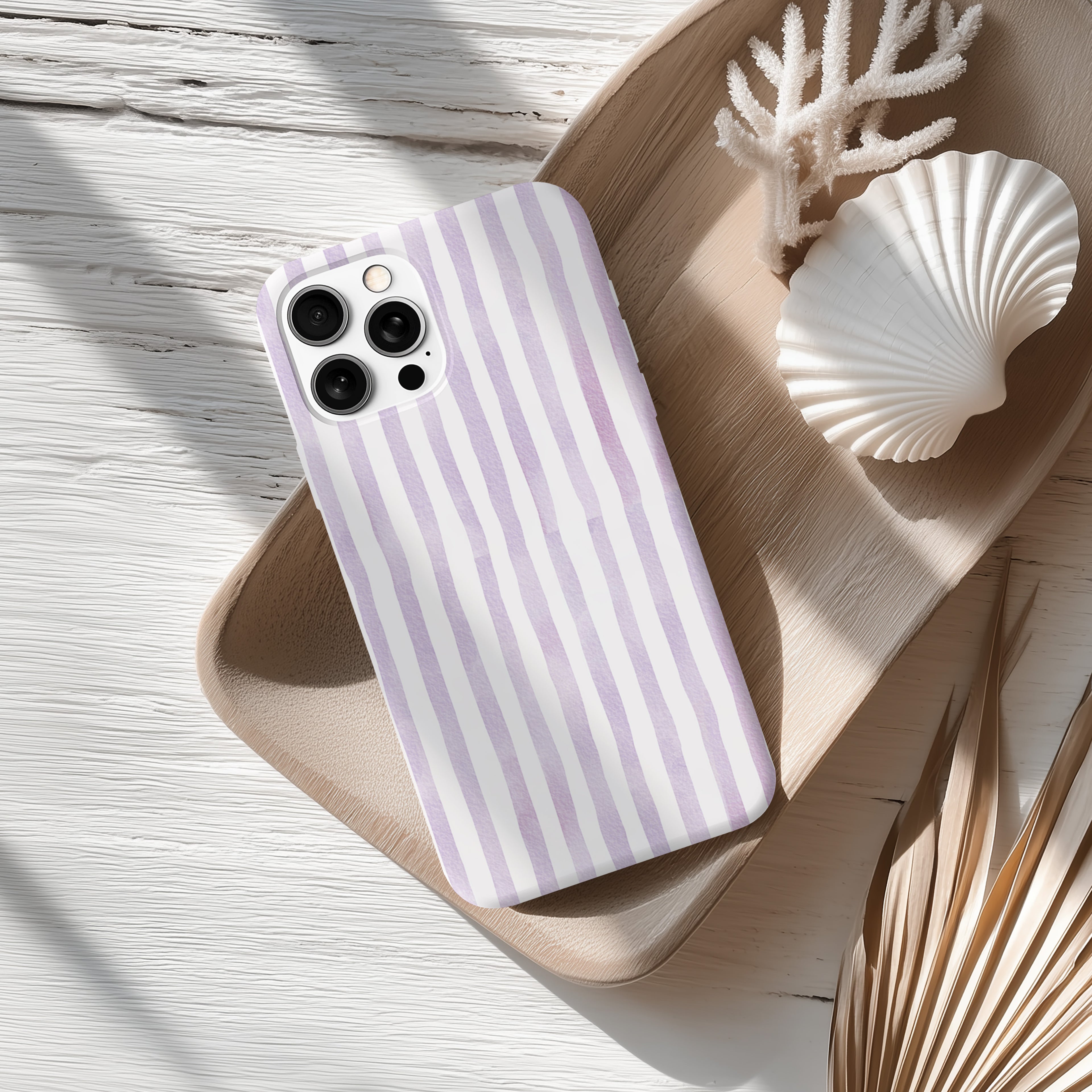A purple and white striped phone case rests on a wooden surface, accompanied by a seashell and some dried leaves.