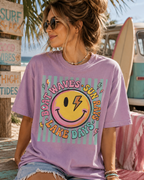 A woman wearing a purple t-shirt with a smiling smiley face and text that says "Surf, Shacks, Good Vibes, Boat Waves, Sun Rays, Lake Days" is sitting on a bench near a beach.