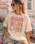 A woman wearing a t-shirt with the text "Boat Sun Lake" and sunglasses is sitting in front of a beach sign.