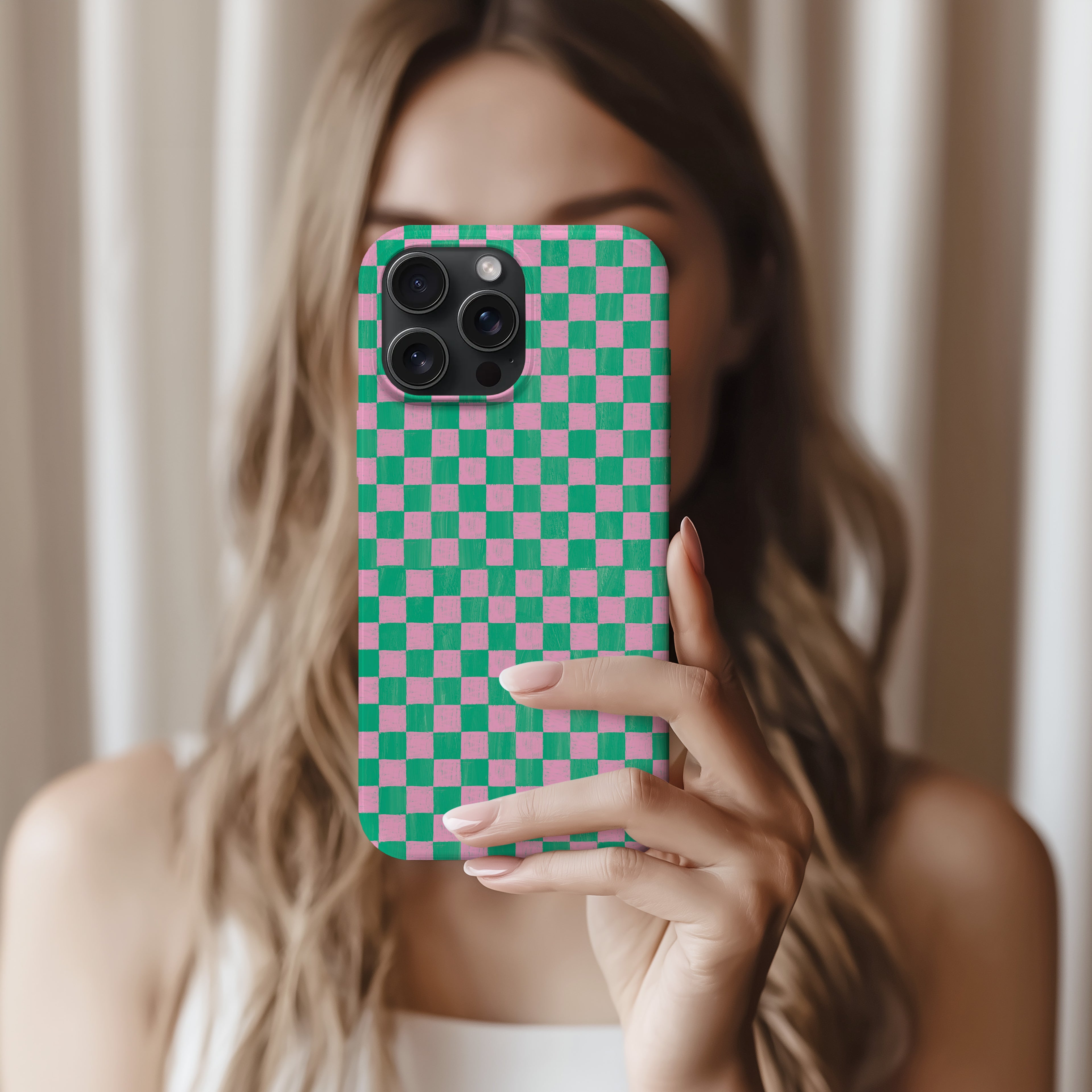 A woman with long, wavy blonde hair is holding a phone with a checkered pattern on it, taking a selfie.
