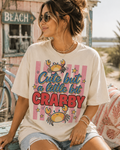A woman wearing a white t-shirt with a crab design and text, sitting on a bench near a beach sign.