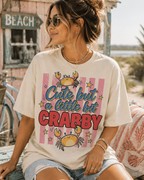A woman wearing a white t-shirt with a crab design and text, sitting on a bench near a beach sign.