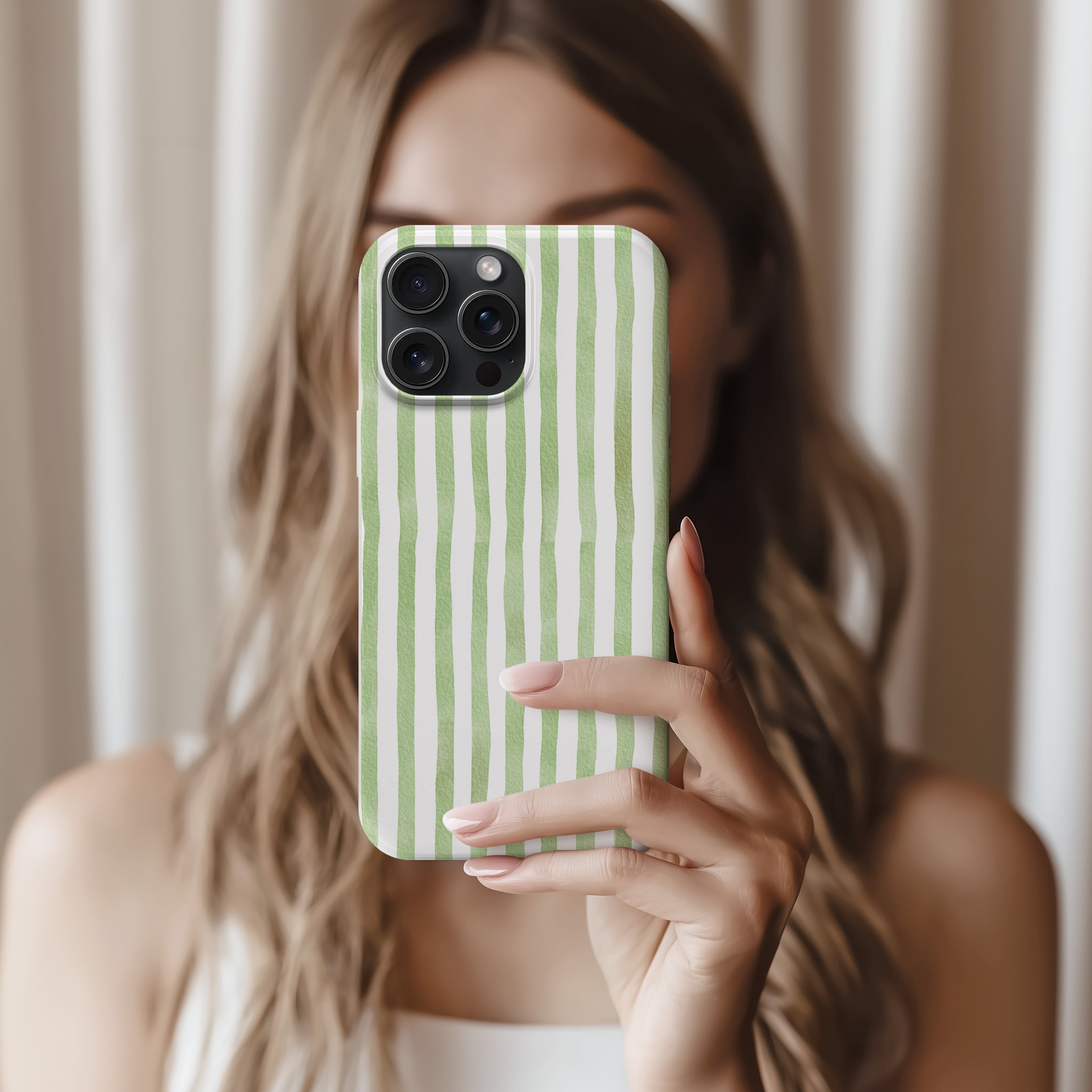 A woman with long, wavy hair is holding a green and white striped phone case up to her face, taking a selfie.