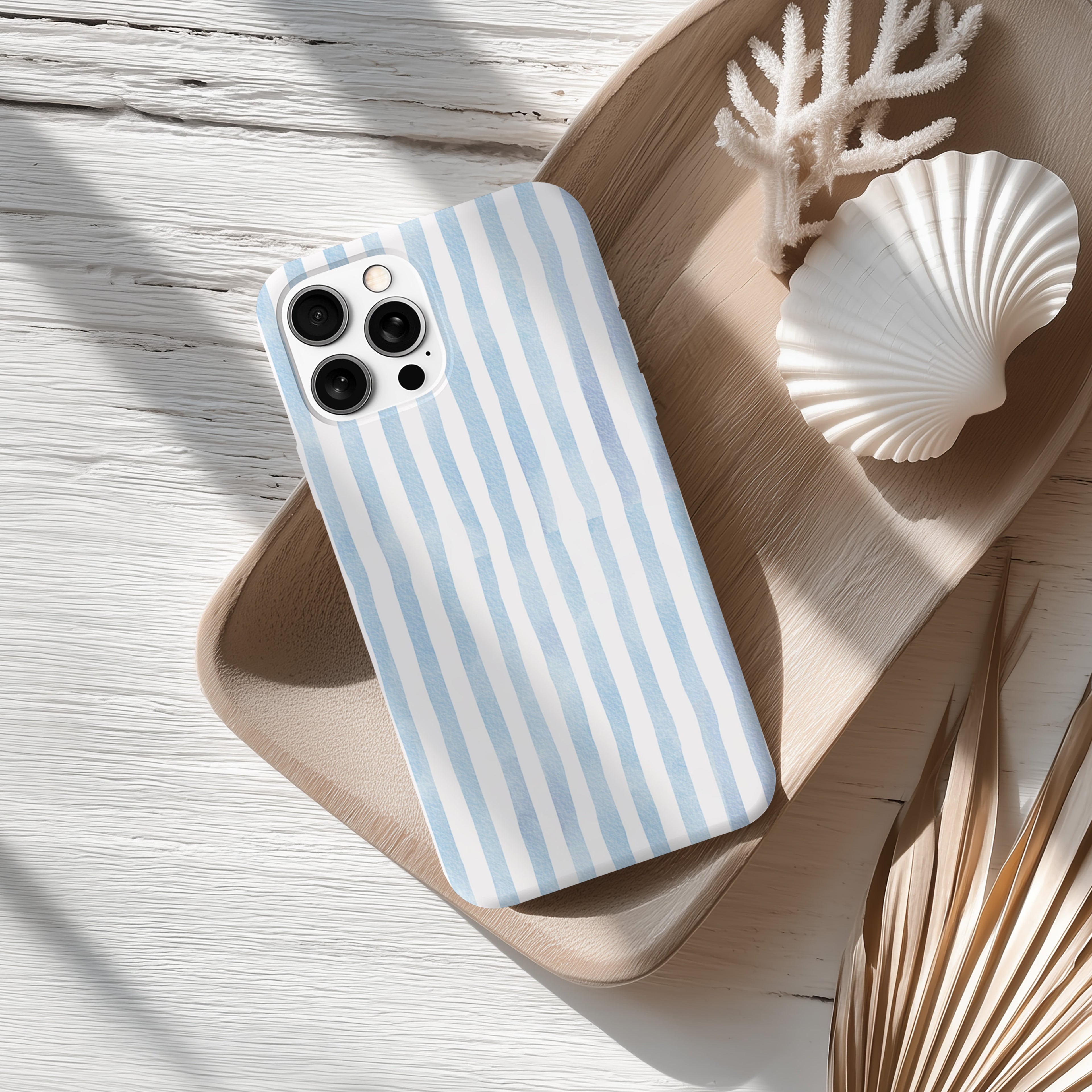 A blue and white striped phone case rests on a wooden surface, accompanied by a white seashell and some dried leaves.