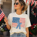 A woman wearing a white t-shirt with an American flag design and holding an American flag is standing in front of a house decorated with American flags.