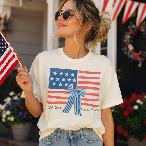 A woman wearing a white t-shirt with an American flag design and holding an American flag is standing in front of a house decorated with American flags.