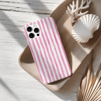 A pink and white striped phone case is placed on a wooden surface, accompanied by a white seashell and some dried leaves.