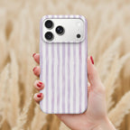 A hand holding a smartphone with a purple and white striped pattern on the back cover, against a blurred background of a field.