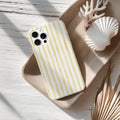 A yellow and white striped phone case rests on a wooden surface, accompanied by a seashell and some dried leaves.