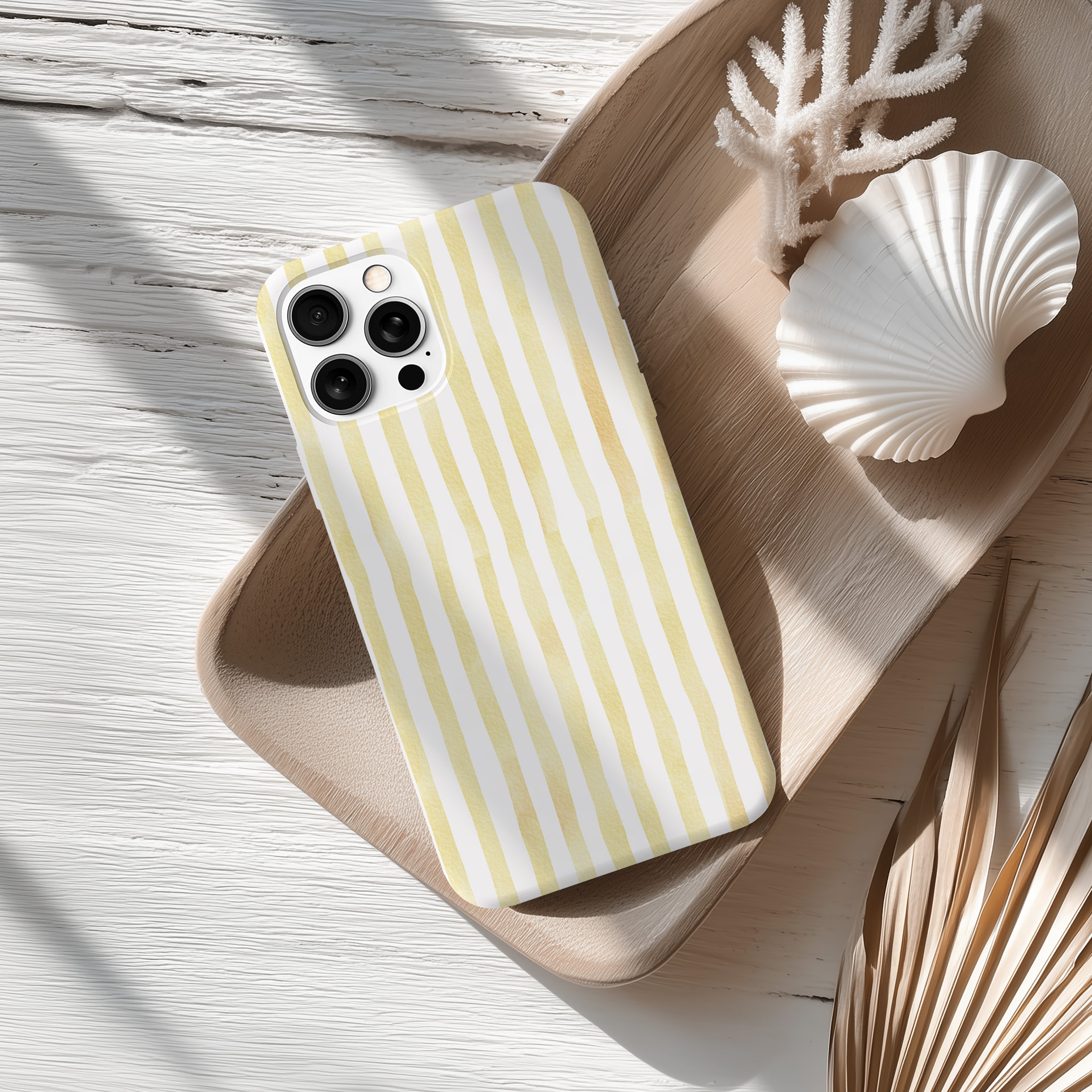 A yellow and white striped phone case rests on a wooden surface, accompanied by a seashell and some dried leaves.